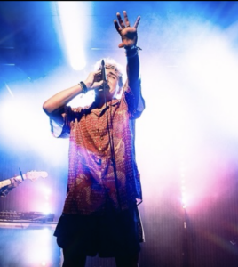 A singer in a patterned shirt performs on stage, proudly representing the LGBTQ community, holding a microphone and raising one hand, with bright stage lights and smoke in the background.